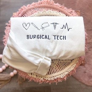 May include: A folded white long-sleeve sweatshirt with black embroidered medical symbols and the text "SURGICAL TECH". The design includes a heart, scalpel, surgical cap, caduceus, and a heartbeat line.
