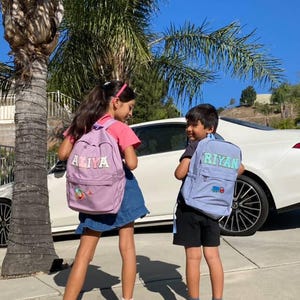May include: Two children with backpacks stand on a pavement. The lavender backpacks have the names "ALIYA" and "RIYAN" in colourful letters. The girl wears a pink top and denim skirt, while the boy wears a black top and shorts. A white car is in the background.