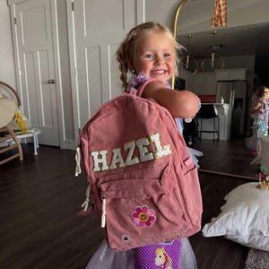May include: A pink corduroy backpack with the name "HAZEL" in white letters. The backpack features a flower patch and white drawstrings. A small purple beaded purse with a unicorn design is also visible.