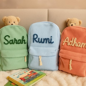 Peut inclure: Trois sacs à dos en velours côtelé vert, bleu et orange, chacun avec un nom brodé sur le devant. Un livre pour enfants avec une illustration de train est visible devant les sacs à dos. Un ours en peluche est posé au-dessus de chaque sac à dos.