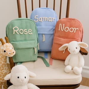 May include: Three corduroy backpacks in green, blue, and coral, each with a name embroidered in white. The backpacks are on a wooden chair with stuffed animal toys. The names on the backpacks are Rose, Samar, and Noor.