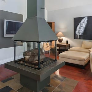 Puede incluir: Una chimenea gris moderna con un cerramiento de vidrio, que muestra troncos y piedras. La chimenea está sobre una base de baldosas, con una chimenea gris a juego. Un televisor de pantalla plana está montado en la pared y una pintura de una pluma cuelga cerca.