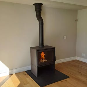 May include: A contemporary black fireplace with a curved flue pipe. The fireplace features a glass door, showcasing a burning fire. It rests on a black base, with a wooden floor in the foreground. The room has light grey walls.