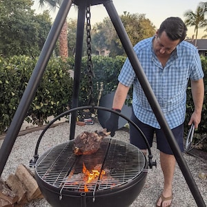 May include: A black metal Cowboy Cauldron tripod grill featuring a suspended, bowl-shaped fire pit with a circular cooking grate. The structure includes a heavy-duty chain for height adjustment and a prominent logo plate at the top apex.