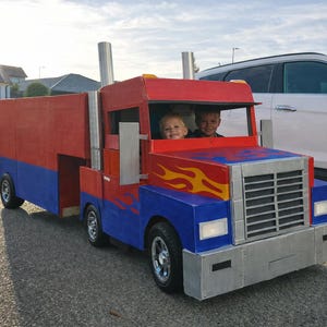 Puede incluir: Un camión de juguete hecho a mano, colorido en rojo, azul y plateado, con detalles de llamas amarillas. Dos niños son visibles dentro de la cabina. El camión tiene un diseño cuadrado con una gran parrilla delantera y tubos de escape.