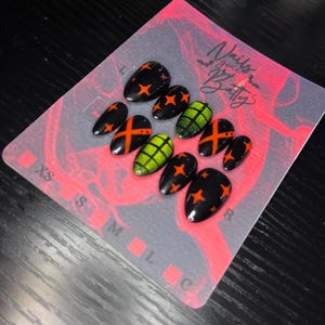 May include: A set of black press-on nails with orange star and cross designs, and green nails with black cross designs. The nails are arranged on a pink and gray card with the text "Nails gone Batty".