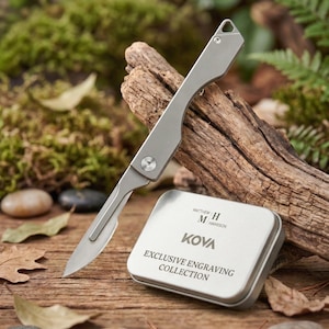 May include: A silver scalpel with a curved handle and a sharp blade, resting on a piece of wood. A small, silver tin with the text "KOVA EXCLUSIVE ENGRAVING COLLECTION" is next to the scalpel. The background includes wood, leaves, and moss.