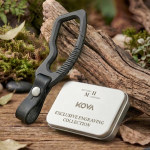 May include: A dark gray multi-tool with a leather strap and a silver snap. A small, silver metal box with the text "KOVA EXCLUSIVE ENGRAVING COLLECTION" is next to the tool. The background includes wood, moss, and small stones.
