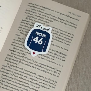 May include: A navy blue sticker in the shape of a jersey with the text "The goal" at the top and "TUCKER 46" in white. A small red heart is at the bottom. The sticker is on an open book.