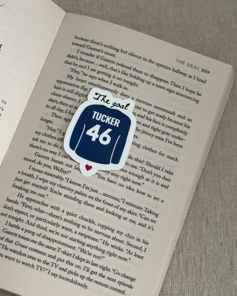 May include: A navy blue sticker in the shape of a jersey with the text "The goal" at the top and "TUCKER 46" in white. A small red heart is at the bottom. The sticker is on an open book.