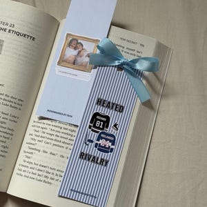 Könnte beinhalten: Ein handgemachtes Lesezeichen mit hellblauem Band, das in ein Buch eingelegt ist. Das Lesezeichen zeigt ein Foto und ein Design mit dem Text "HEATED RIVALRY" und Hockey-Trikot-Grafiken. Das Lesezeichen ist aus Papier.