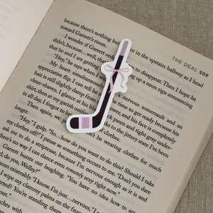 May include: A hockey stick-shaped bookmark with a black and light purple design, accented with a pink bow. The bookmark is placed on an open book, enhancing the visual appeal. A unique accessory for book lovers.