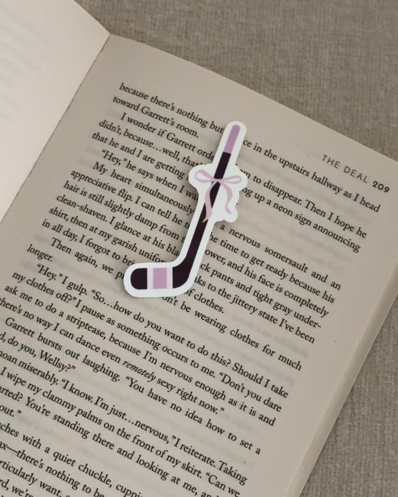 May include: A hockey stick-shaped bookmark with a black and light purple design, accented with a pink bow. The bookmark is placed on an open book, enhancing the visual appeal. A unique accessory for book lovers.
