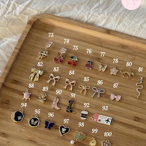Puede incluir: Una colecci&oacute;n de peque&ntilde;os dijes decorativos de varias formas y colores, incluyendo lazos, cerezas, mariposas y botas. Los dijes est&aacute;n dispuestos en una bandeja de madera, con n&uacute;meros.