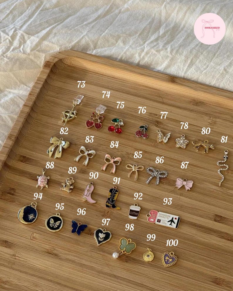 Puede incluir: Una colecci&oacute;n de peque&ntilde;os dijes decorativos de varias formas y colores, incluyendo lazos, cerezas, mariposas y botas. Los dijes est&aacute;n dispuestos en una bandeja de madera, con n&uacute;meros.