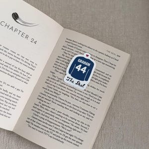May include: A bookmark featuring a navy blue jersey design with the name "GRAHAM" and the number "44" in white. The text "The Deal" is below the jersey. The bookmark rests on an open book.