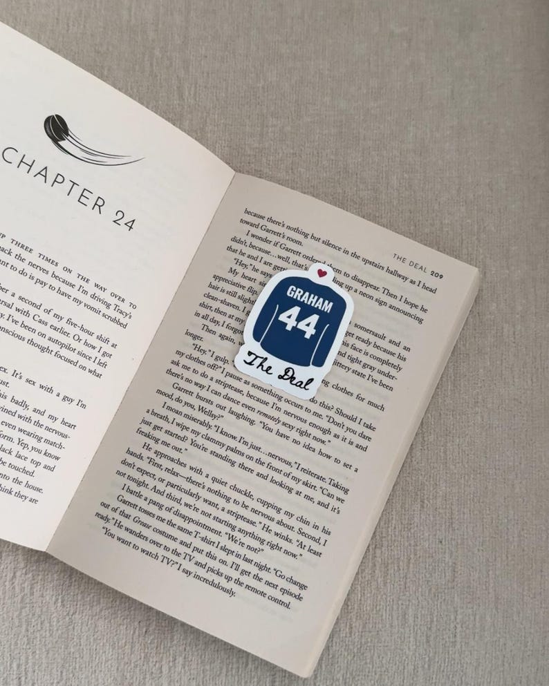 May include: A bookmark featuring a navy blue jersey design with the name "GRAHAM" and the number "44" in white. The text "The Deal" is below the jersey. The bookmark rests on an open book.