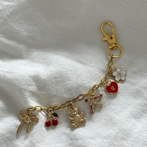 May include: Gold charm bracelet with a variety of charms, including a teddy bear, a heart with a keyhole, a bow, a cherry, and a flower. The bracelet has a gold lobster clasp.