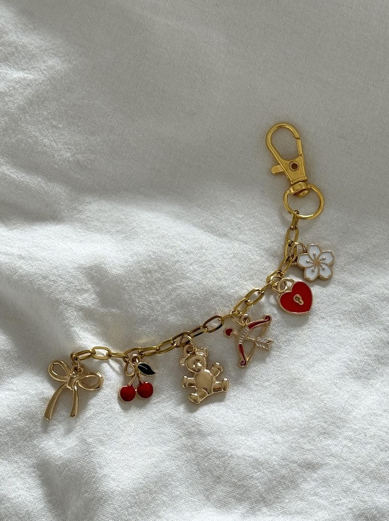 May include: Gold charm bracelet with a variety of charms, including a teddy bear, a heart with a keyhole, a bow, a cherry, and a flower. The bracelet has a gold lobster clasp.