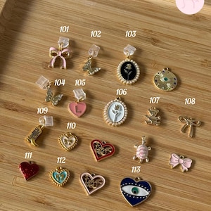Puede incluir: Una colecci&oacute;n de dijes dorados con varios dise&ntilde;os, incluyendo lazos, mariposas, corazones y una tortuga. Algunos dijes est&aacute;n acentuados con esmalte rosa, rojo y azul. Los dijes se exhiben sobre una superficie de madera, cada uno etiquetado con un n&uacute;mero.