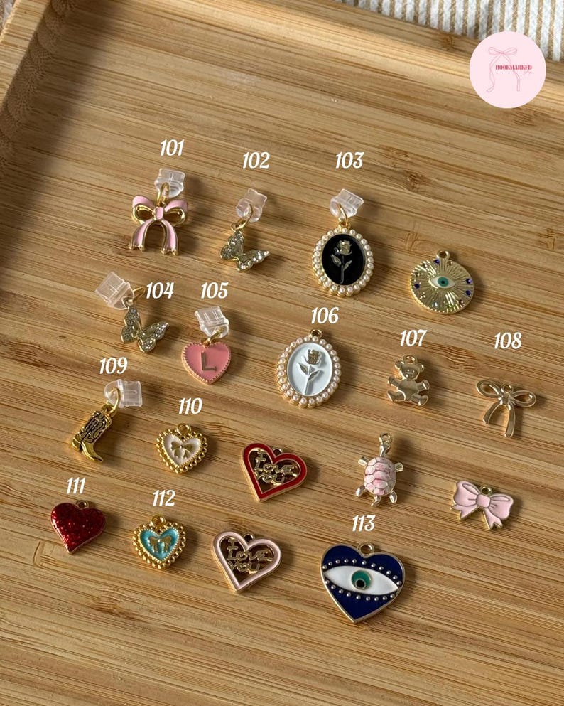 Puede incluir: Una colecci&oacute;n de dijes dorados con varios dise&ntilde;os, incluyendo lazos, mariposas, corazones y una tortuga. Algunos dijes est&aacute;n acentuados con esmalte rosa, rojo y azul. Los dijes se exhiben sobre una superficie de madera, cada uno etiquetado con un n&uacute;mero.