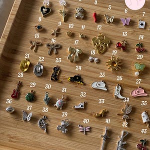 Puede incluir: Una colecci&oacute;n de peque&ntilde;os dijes decorativos en varias formas, incluyendo un coraz&oacute;n, un oso de peluche, una mariposa, un sombrero de vaquero, y m&aacute;s. Los dijes est&aacute;n dispuestos en una bandeja de madera, mostrando una variedad de dise&ntilde;os y colores.