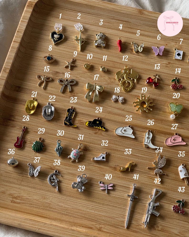 Puede incluir: Una colecci&oacute;n de peque&ntilde;os dijes decorativos en varias formas, incluyendo un coraz&oacute;n, un oso de peluche, una mariposa, un sombrero de vaquero, y m&aacute;s. Los dijes est&aacute;n dispuestos en una bandeja de madera, mostrando una variedad de dise&ntilde;os y colores.