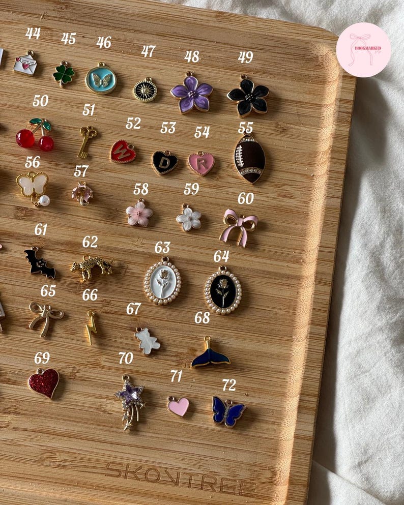 Puede incluir: Una colecci&oacute;n de dijes dorados con varios dise&ntilde;os, incluyendo flores, corazones, mariposas y un bal&oacute;n de f&uacute;tbol americano. Los dijes est&aacute;n dispuestos en una bandeja de madera, mostrando una variedad de colores y formas. La bandeja tiene la palabra "SKONTREE" impresa.
