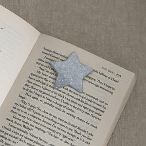 May include: A glittery, silver star-shaped bookmark rests on the open pages of a book. The bookmark is made of a sparkly material and is positioned near the top of the page. The book's text is visible, with the title "THE DEAL" on the top right.