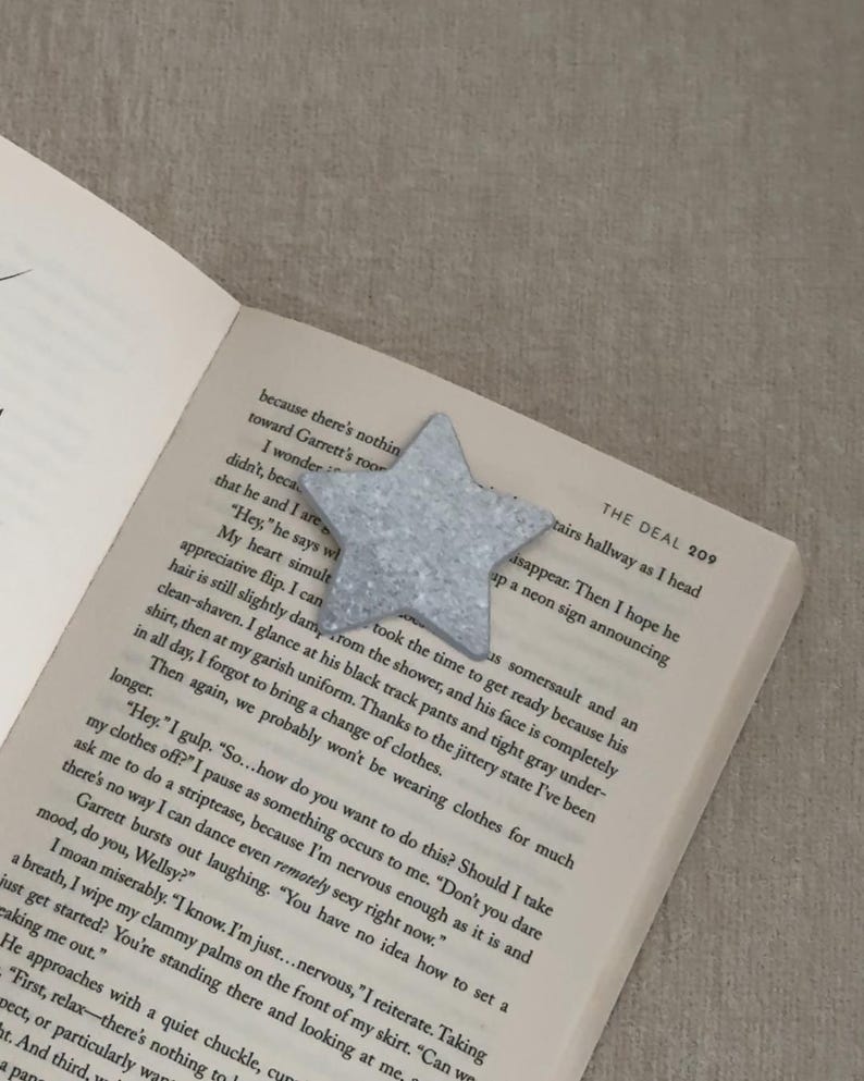 May include: A glittery, silver star-shaped bookmark rests on the open pages of a book. The bookmark is made of a sparkly material and is positioned near the top of the page. The book's text is visible, with the title "THE DEAL" on the top right.
