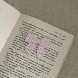 May include: A light pink plaid bow-shaped bookmark rests on the open pages of a book. The bookmark is made of paper and has a simple, elegant design. The book's text is visible beneath the bookmark.