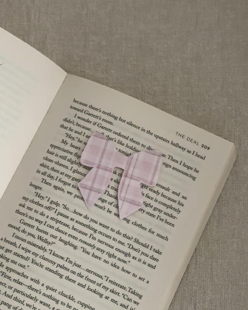 May include: A light pink plaid bow-shaped bookmark rests on the open pages of a book. The bookmark is made of paper and has a simple, elegant design. The book's text is visible beneath the bookmark.