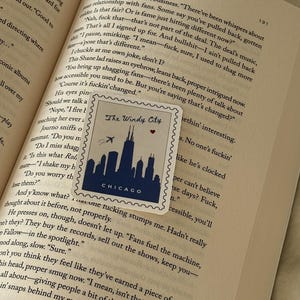 May include: A navy blue sticker with a white border featuring a Chicago skyline silhouette, the text "The Windy City" and a small red heart. The sticker is placed on an open book.