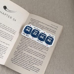 May include: A bookmark featuring jersey-style designs with names and numbers: Graham 44, Logan 22, Di Laurentis 66, and Tucker 46. The bookmark is placed on an open book with text.