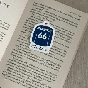 May include: A navy blue and white sticker featuring a jersey design with the text "DI LAURENTIS 66" and "The Score". The sticker is placed on an open book with printed text.