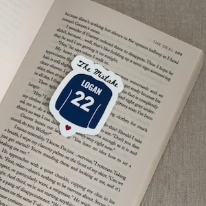May include: A navy blue sticker in the shape of a jersey with the text "The Mistake", "LOGAN", and the number "22". A small red heart is at the bottom. The sticker is on top of an open book.