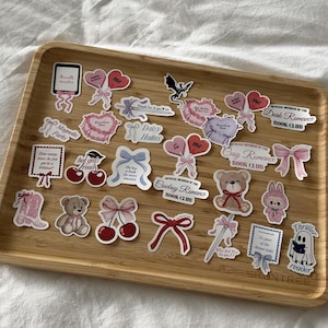 Könnte beinhalten: Eine Holzablage mit Aufklebern. Die Aufkleber zeigen verschiedene Designs, darunter Herzen, Schleifen, Kirschen, einen Teddybären und Texte wie "Kindle reader", "Dark Romance Book Club" und "Thriller reader". Die Aufkleber sind in Rosa-, Rot- und Weißtönen gehalten.