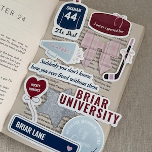 May include: A collection of stickers on an open book. Stickers include a hockey jersey with "GRAHAM 44", a hockey puck, a hockey stick, and text that reads "BRIAR UNIVERSITY" and "BRIAR LANE". The stickers are in shades of blue, burgundy, and white.