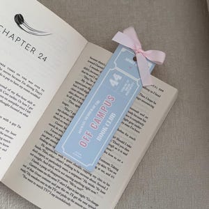 May include: A light blue bookmark with the text "OFF CAMPUS BOOK CLUB" and "44" in pink. The bookmark is tied with a pink ribbon and is placed in an open book.