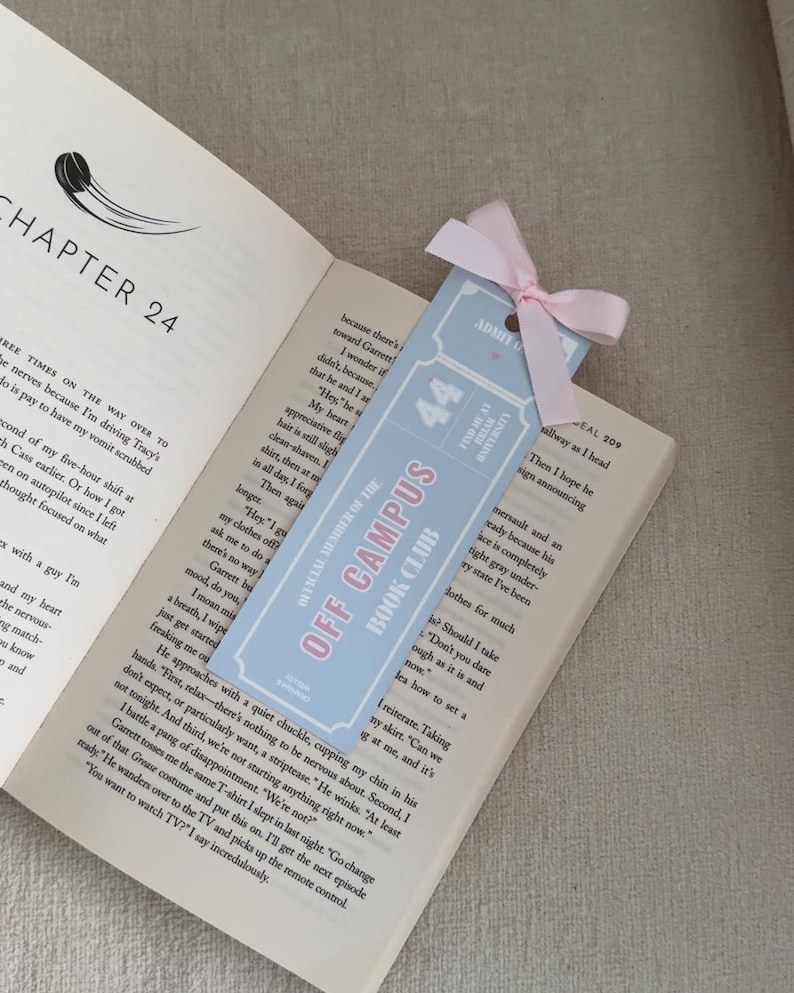 May include: A light blue bookmark with the text "OFF CAMPUS BOOK CLUB" and "44" in pink. The bookmark is tied with a pink ribbon and is placed in an open book.