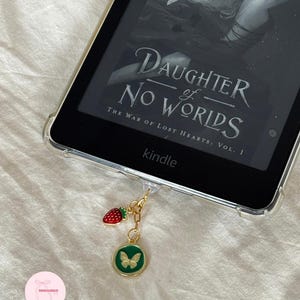 Puede incluir: Un lector de libros electr&oacute;nicos Kindle con una funda protectora transparente que muestra el texto "DAUGHTER of NO WORLDS". Un dije dorado con una fresa roja y un colgante de mariposa verde cuelga del dispositivo. El lector electr&oacute;nico est&aacute; sobre una superficie blanca.