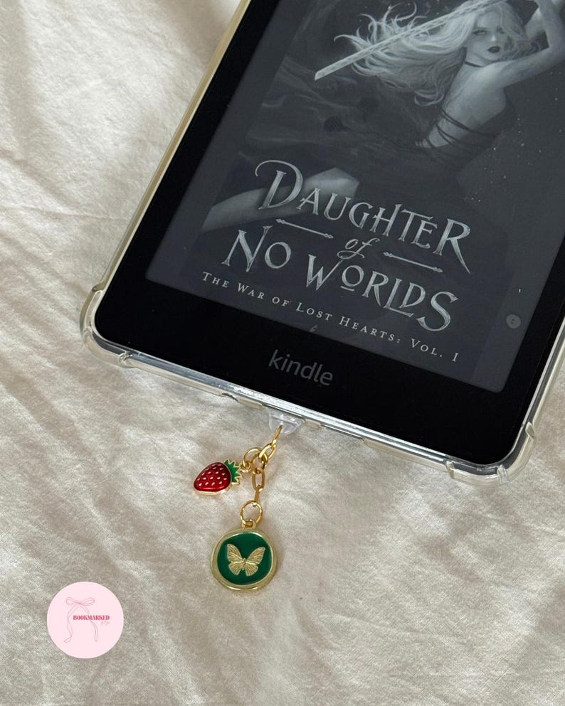 Puede incluir: Un lector de libros electr&oacute;nicos Kindle con una funda protectora transparente que muestra el texto "DAUGHTER of NO WORLDS". Un dije dorado con una fresa roja y un colgante de mariposa verde cuelga del dispositivo. El lector electr&oacute;nico est&aacute; sobre una superficie blanca.