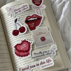 Pode incluir: Uma coleção de autocolantes com temas românticos, colocados num livro aberto. Os autocolantes apresentam frases como "Always falling for the villain" e "I need you in this life". Outros autocolantes retratam cerejas, lábios e um design em forma de coração.