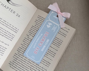 Op de afbeelding: Een lichtblauwe bladwijzer met de tekst "OFF CAMPUS BOOK CLUB" en "44" in roze. De bladwijzer is vastgebonden met een roze lint en ligt in een open boek.