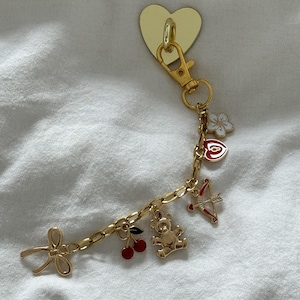 May include: A gold charm bracelet with a heart-shaped charm, a bow charm, a teddy bear charm, a cherry charm, and an arrow charm.