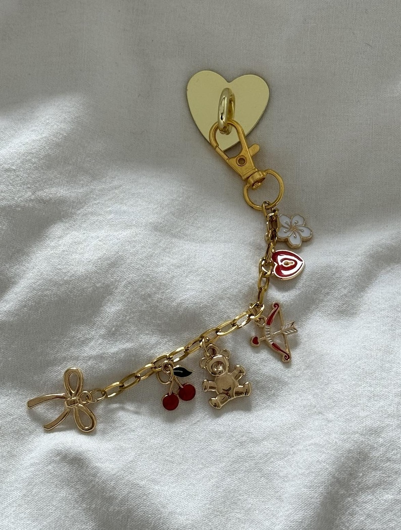 May include: A gold charm bracelet with a heart-shaped charm, a bow charm, a teddy bear charm, a cherry charm, and an arrow charm.