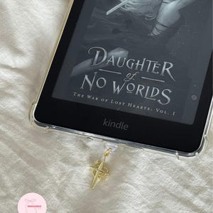 Puede incluir: Un lector de libros electr&oacute;nicos Kindle con una funda protectora transparente que muestra la portada del libro "Daughter of No Worlds". Un dije dorado en forma de br&uacute;jula cuelga del dispositivo. El fondo es una superficie blanca texturizada.