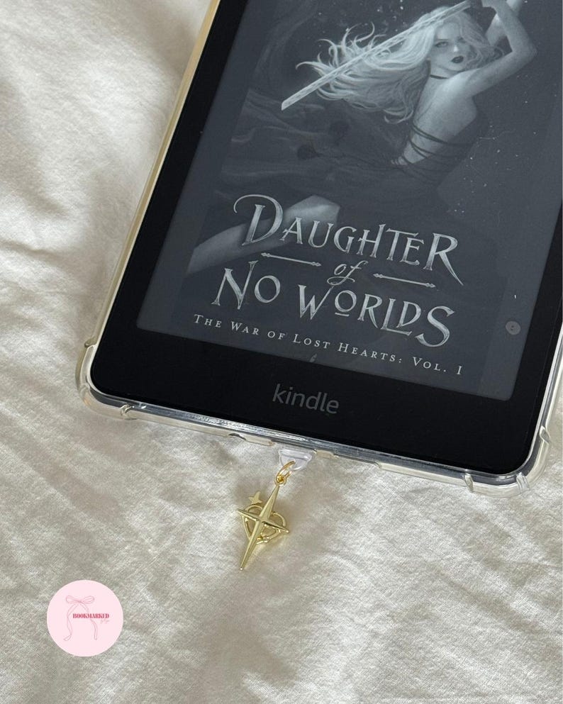 Puede incluir: Un lector de libros electr&oacute;nicos Kindle con una funda protectora transparente que muestra la portada del libro "Daughter of No Worlds". Un dije dorado en forma de br&uacute;jula cuelga del dispositivo. El fondo es una superficie blanca texturizada.