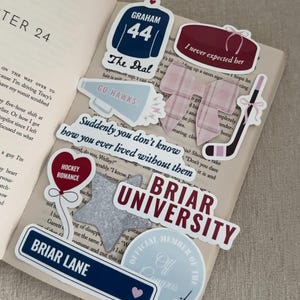 May include: A collection of stickers on an open book. Stickers include a hockey jersey, a hockey puck, a hockey stick, and text that reads "Briar University", "Briar Lane", and "Hockey Romance".