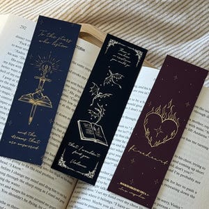 Può includere: Tre segnalibri decorativi con disegni in lamina d'oro. Uno è blu navy con una spada e testo, un altro è nero con un libro e draghi, e l'ultimo è bordeaux con un cuore e fiamme. Tutti hanno bordi decorativi.